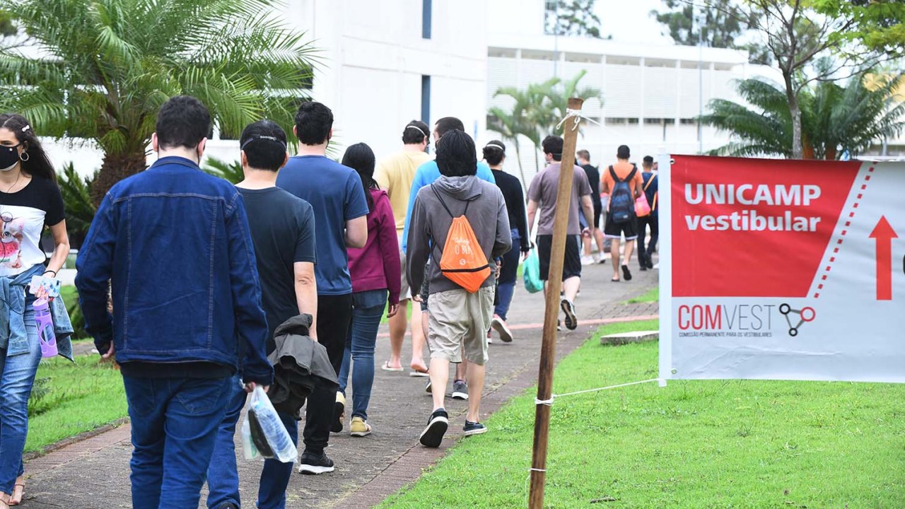 Faixa indica com uma seta vermelha caminho para as salas de aplicação do vestibular da Unicamp. Um grupo de estudantes, de costas para a câmera, anda em direção a um prédio.