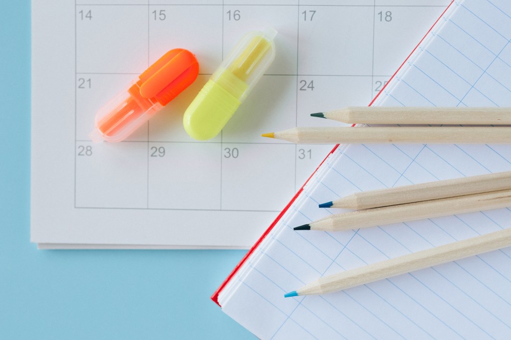 Mesa azul com calendário, lápis coloridos e caderno pautado aberto.