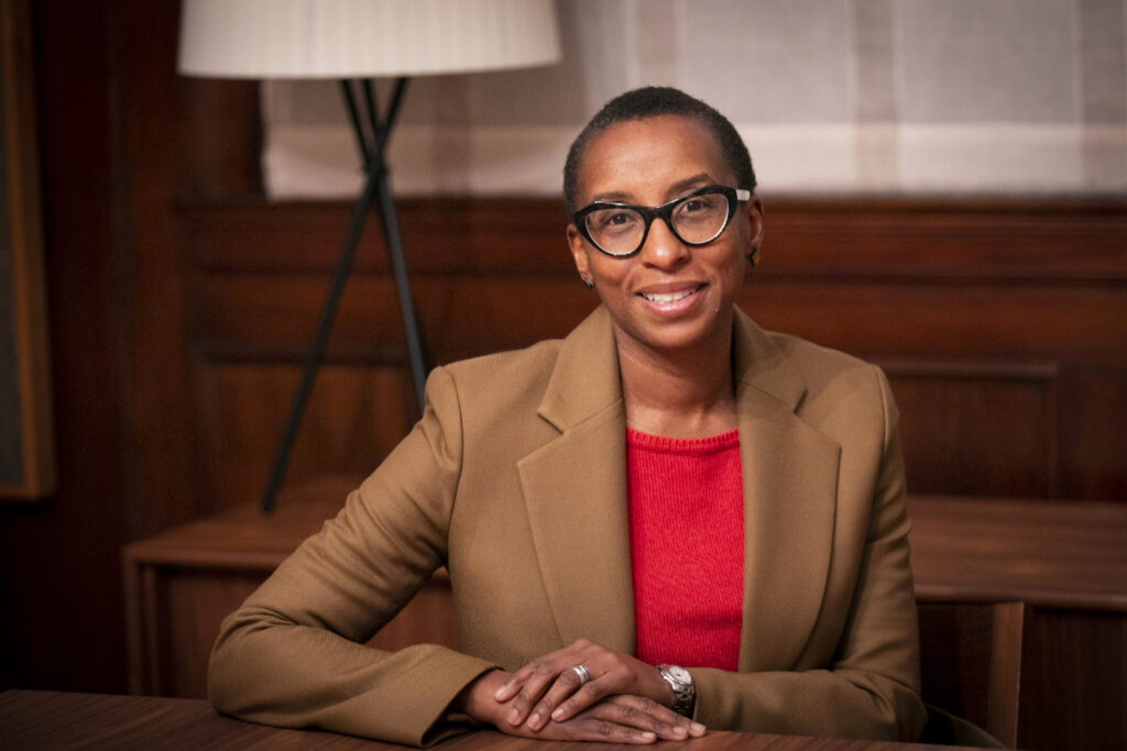 A presidente da Universidade de Harvard, Claudine Gay. Ela é uma mulher negra, de óculos de grau, e cabelo raspado. Usa um terno bege, e está sentada com os braços cruzados sobre uma mesa.