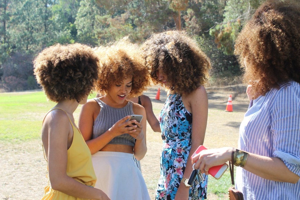 Grupo de mulheres conversando e checando o celular. Todas são negras, de cabelo afro, apresentam estarem felizes e estão em um parque.