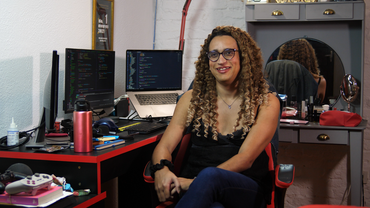 Estudante Nathally Souza sentada em frente a uma mesa, com dois computadores. Ela é negra, usa cabelo trançado e usa óculos de grau.