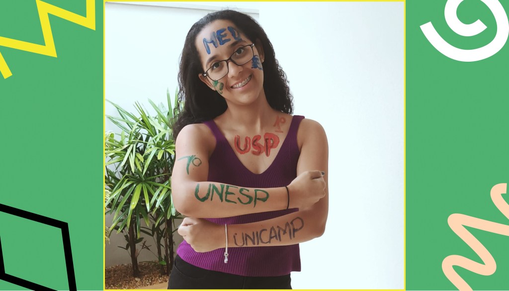 Gabriella, com uma blusa roxa e óculos pretos, mostra os braços com os nomes das universidades: Unicamp e Unesp. Ainda tem pintado com tintas coloridas no seu busto "USP"e "MED"