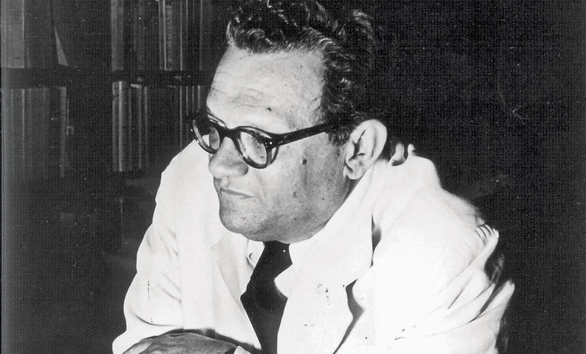 O autor José Lins do Rego é um homem branco, de cabelo escuro e usa óculos. Está sentado, com os braços apoiados em cima de uma mesa.