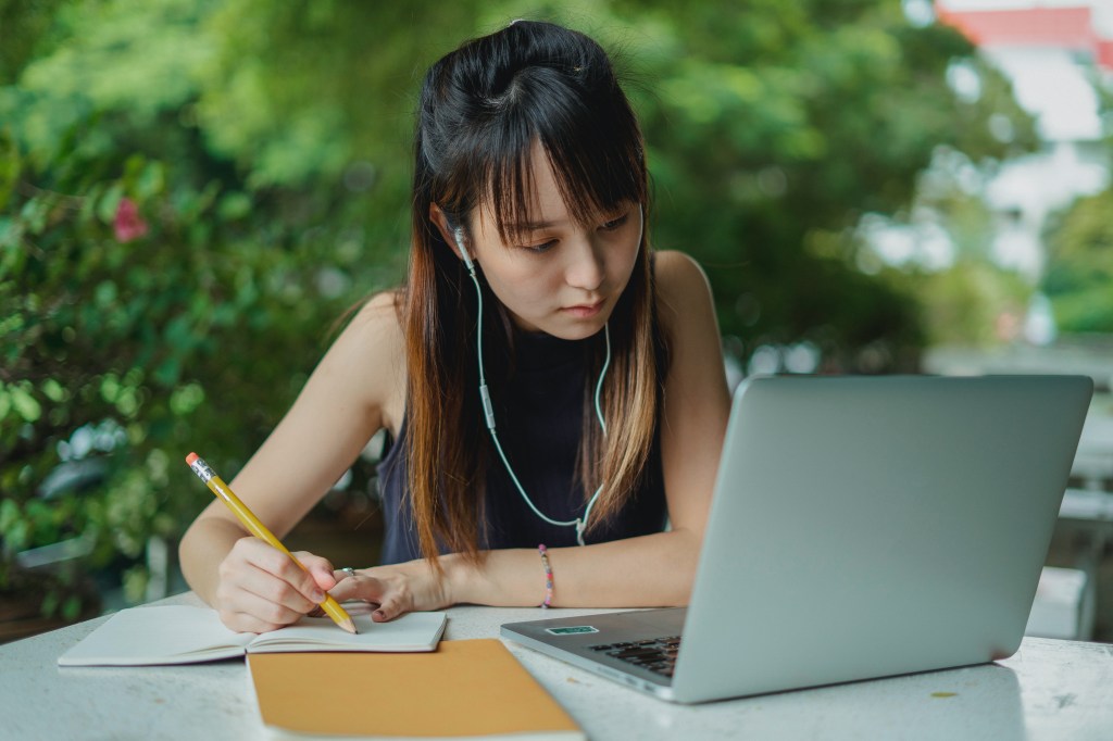 Garota estudando no computador com fone de ouvido, caderno e lápis na mão
