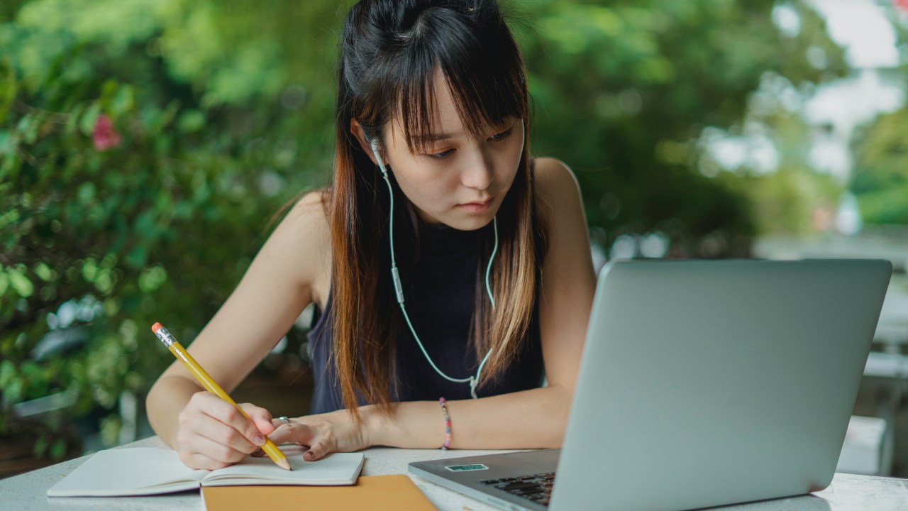 Garota estudando no computador com fone de ouvido, caderno e lápis na mão