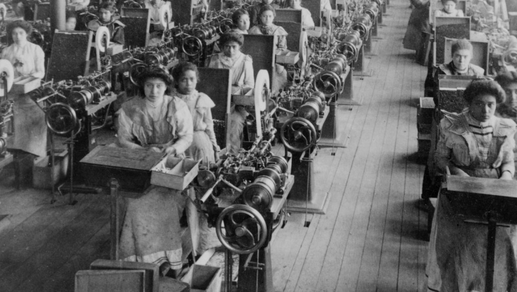 Mulheres trabalhando operando máquinas, elas estão sentadas e olham para a câmera.