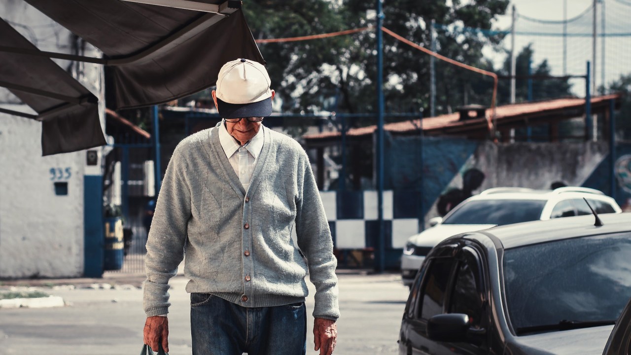 Homem idoso de cabeça baixa anda na calçada com sacola na mão