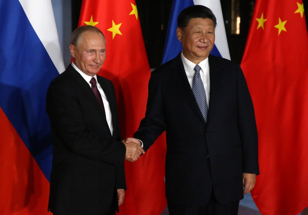 presidente russo vladimir putin aperta a mão do presidente chinês Xi Jinping. Ao fundo, bandeiras dos países-membros do BRICS