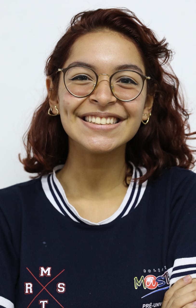 Ana Alice Teixeira Freire é uma mulher de 17 anos. Tem cabelos castanhos ondulados, usa óculos de grau e está com o uniforme do colégio. Ela sorri para a câmera.