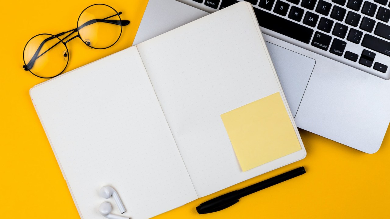 Fotografia mostrar visão superior de uma mesa de cor amarelo vivo, nela estão: um caderno aberto, com as folhas em branco, um óculos de grau, parte de um notebook e um parte de fones de ouvidos sem fio.