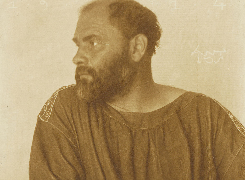 Gustav Klimt foi um homem branco, calvo e barbudo. A fotografia, em cor sépia, o mostra de perfil, usando um traje avermelhado.