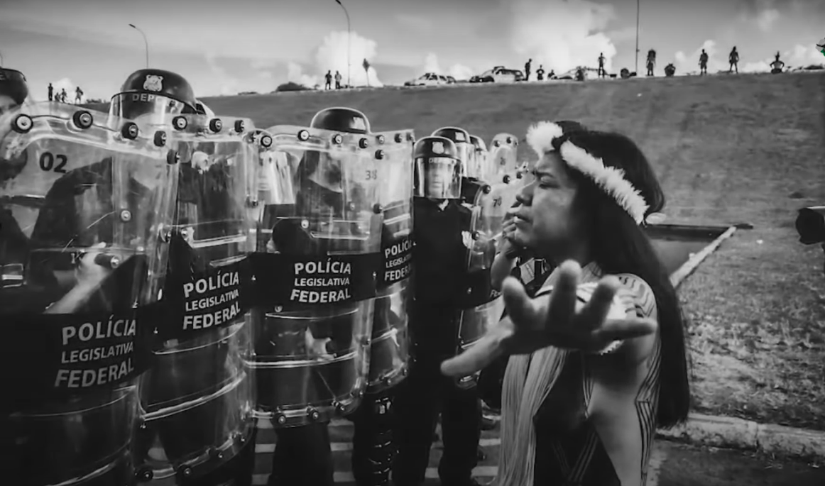 Cena do documentário "Guerras do Brasil.doc" mostra mulher indígena de braços abertos, em frente a batalhão da polícia.