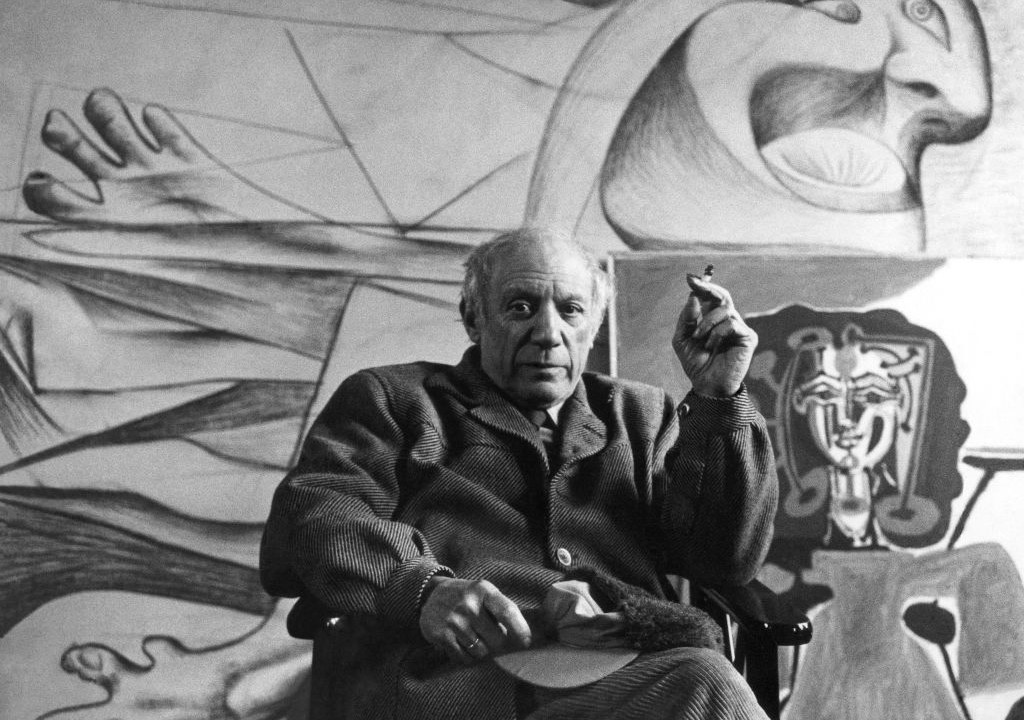 o artista Pablo Picasso sentado em uma poltrona enquanto fuma. Ao fundo, suas obras