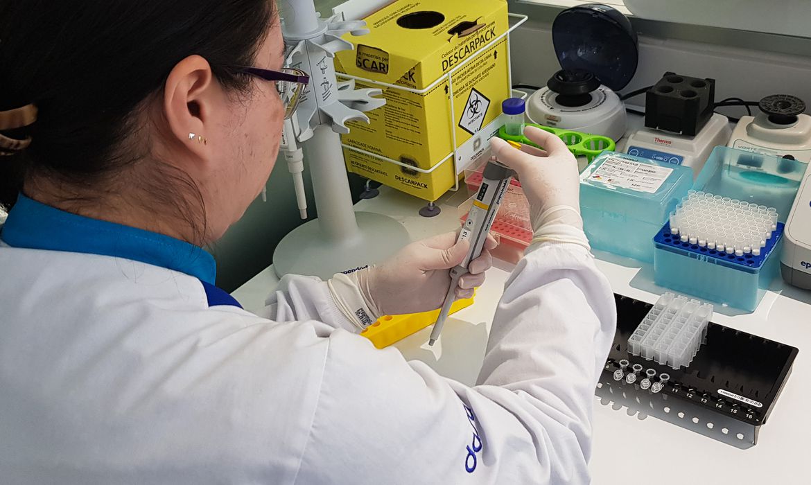Cientista brasileira sentada de costas em procedimento de extração de DNA. Ela é branca, tem cabelos negros presos, usa óculos, avental branco e luvas cirúrgicas.