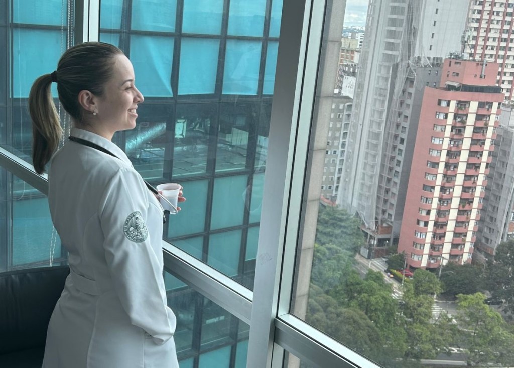 a estudante de medicina bruna, mulher branca de cabelos loiros, veste um jaleco a observa uma paisagem de prédios pela janela