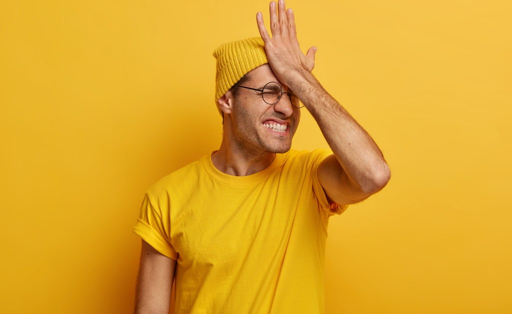 Homem de camiseta amarela, touca e óculos de grau está com semblante preocupado, com a palma do mão direita na testa. Ele parece frustado, irritado.