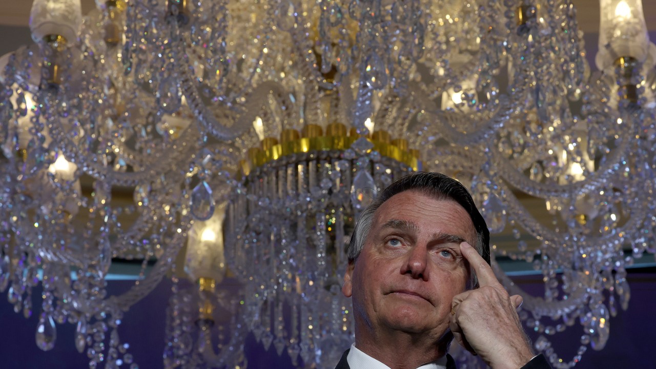 o ex-presidente Jair Bolsonaro, com a mão na sobrancelha, em frente a um lustre luxuoso