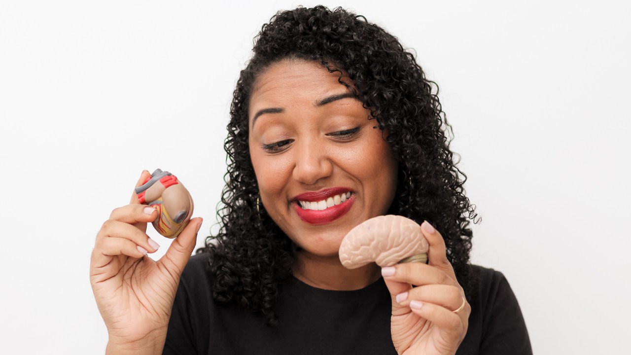 Professora Rafaela, uma mulher negra de pele clara e cabelos cacheados, segura miniaturas representando órgãos humanos