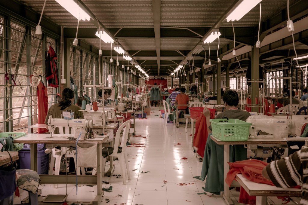 Galpão de produção de roupas com muitos retalhos no chão e mulheres costurando