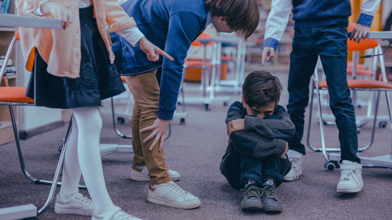 Crianças apontando para um menino encolhido no chão.