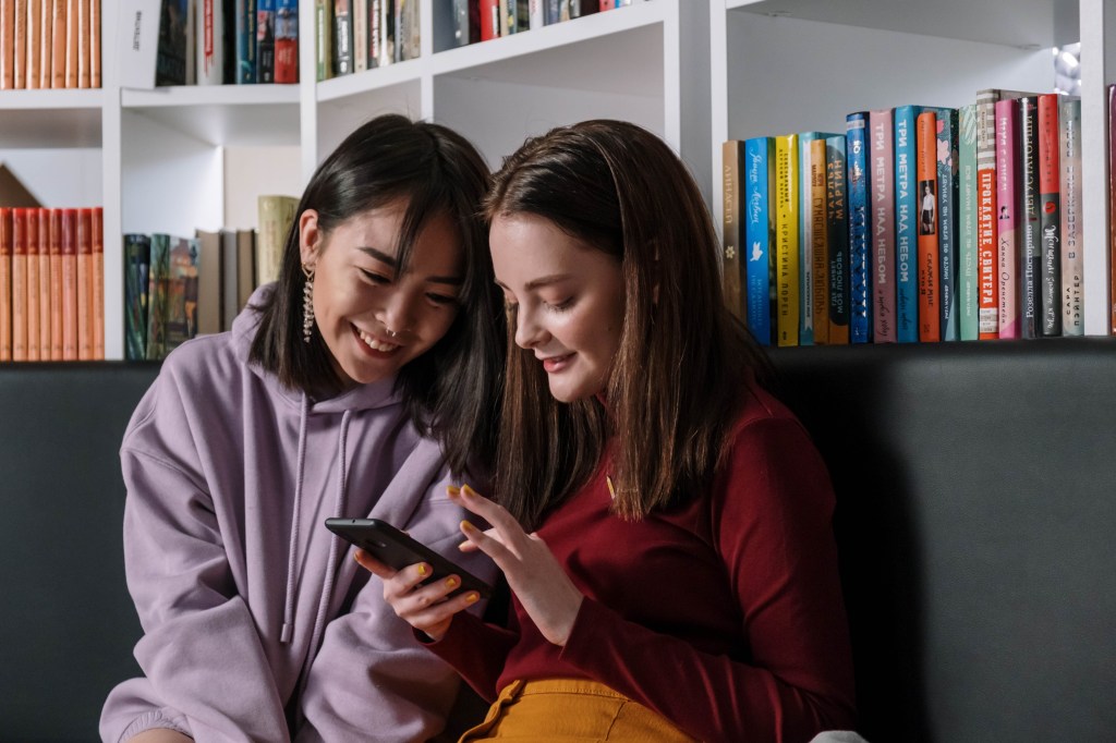 duas meninas mexendo no celular em frente a uma estante de livros