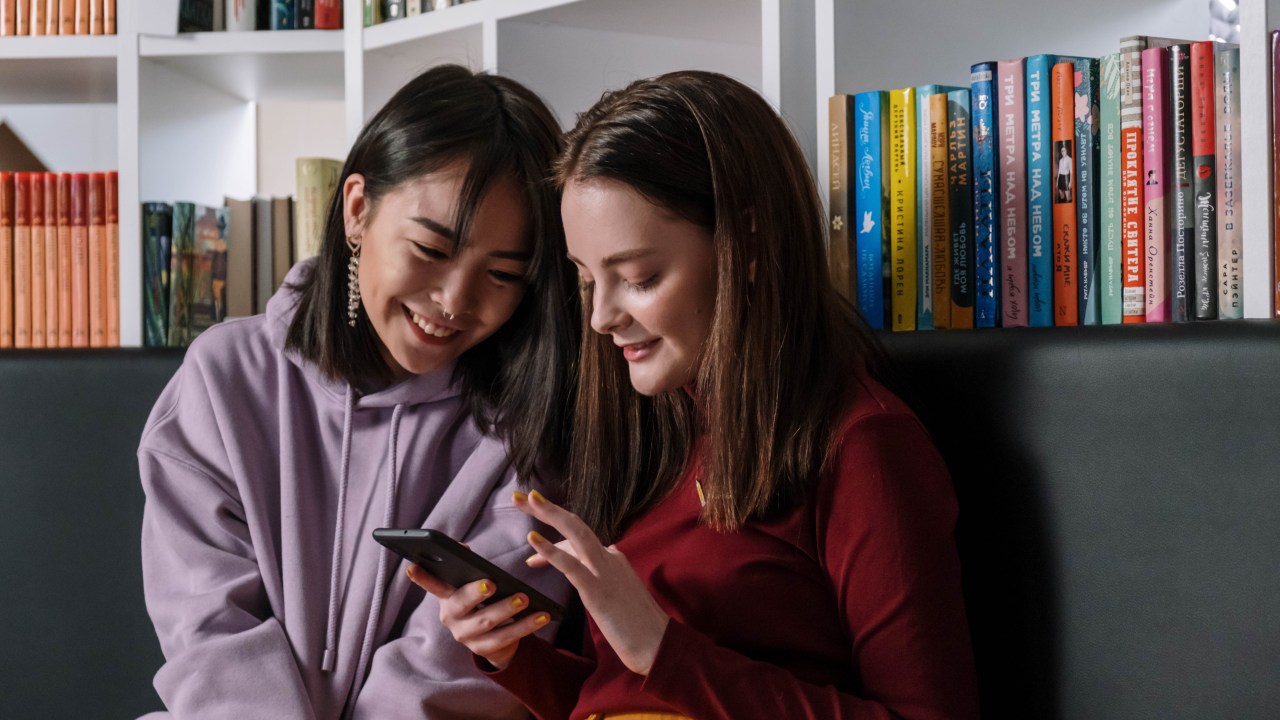 duas meninas mexendo no celular em frente a uma estante de livros