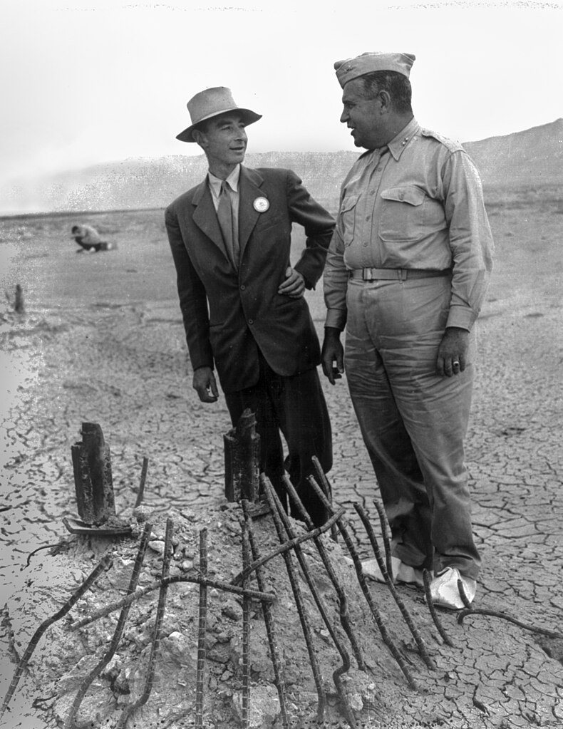 J. Robert Oppenheimer e Leslie Groves analisando os restos da Experiência Trinity em setembro de 1945. As galochas brancas evitavam o derretimento das solas dos seus sapatos.[51]