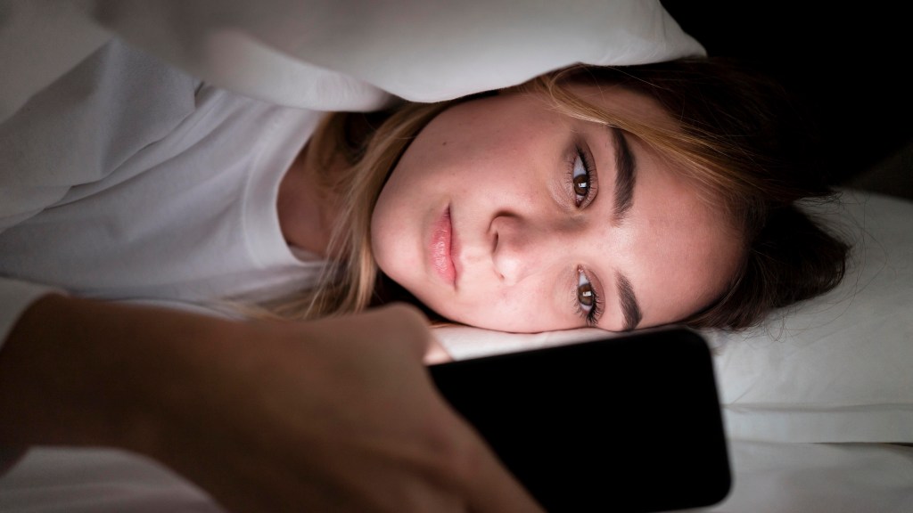 Mulher deitada na cama mexendo no celular próximo ao rosto