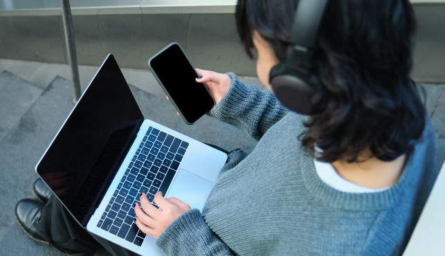 jovem mulher sentada mexendo no notebook, celular e usando fone de ouvido