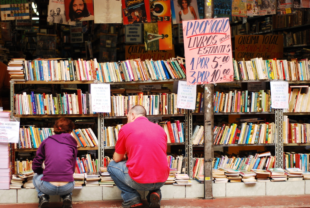 Homem e menina ajoelhado em frente à prateleira com livros em exposição em sebo de São Paulo