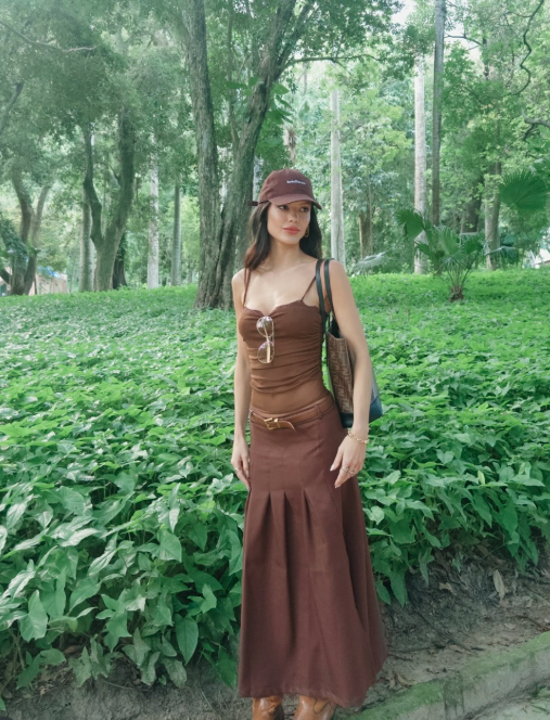 Atriz Vitória Strada. Ela usa um vestido e boné marrons e está em frente a diversas plantas