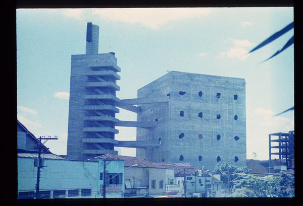Fotografia colorida de um grande edif&iacute;cio em concreto bruto, com torre lateral de circula&ccedil;&atilde;o em escadas abertas e corpo principal retangular perfurado por janelas irregulares, erguido acima de um conjunto de casas baixas em uma &aacute;rea urbana sob c&eacute;u azul com poucas nuvens.