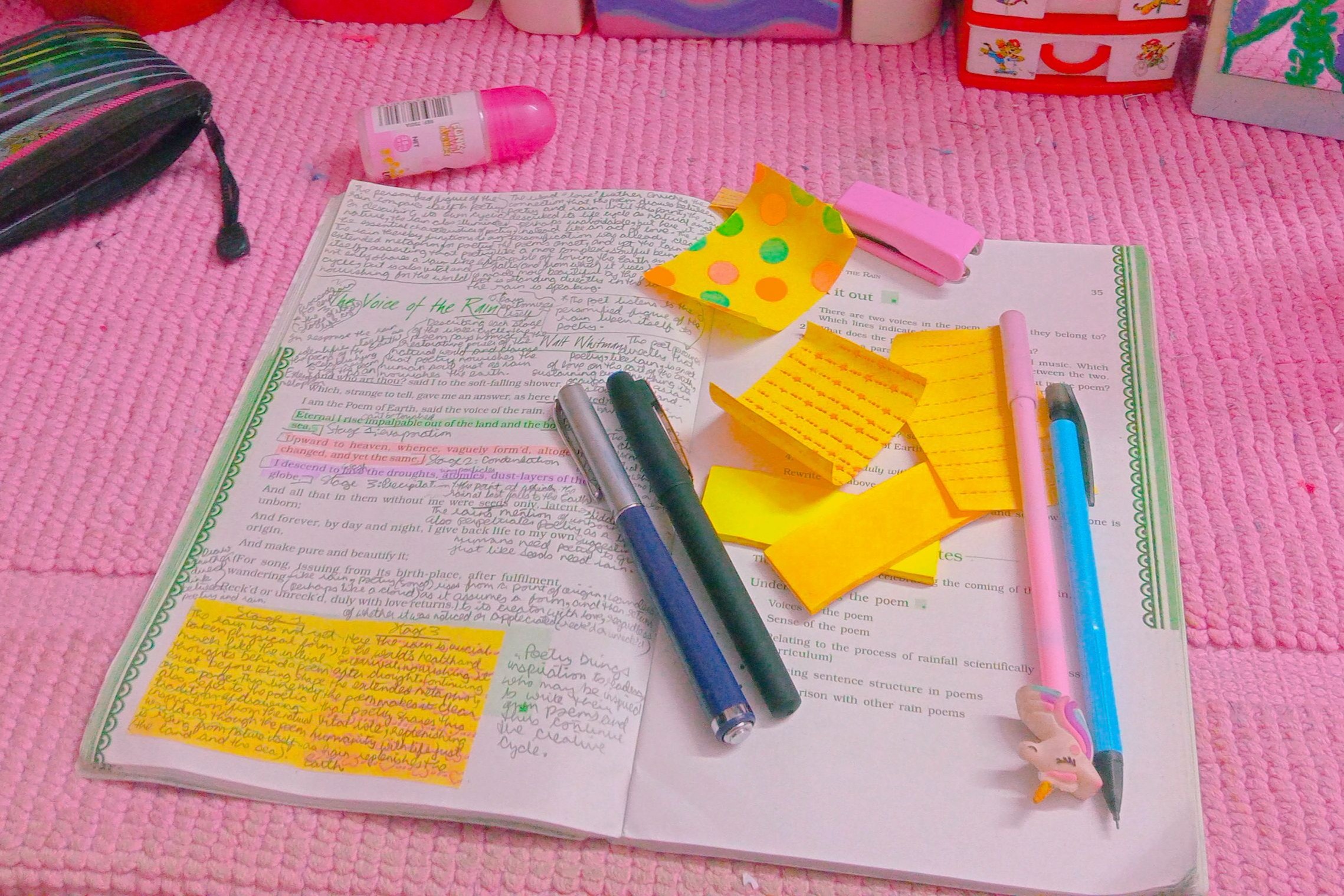Caderno aberto sobre um tapete rosa fofo, repleto de anotações manuscritas detalhadas em várias cores de caneta, com post-its coloridos amarelos, rosa e laranja colados nas páginas e margens. Ao redor, itens de estudo como canetas verde e azul, borracha rosa, estojo, frascos de perfume e caixas decoradas, tudo em estética kawaii com filtro de cor rosado.