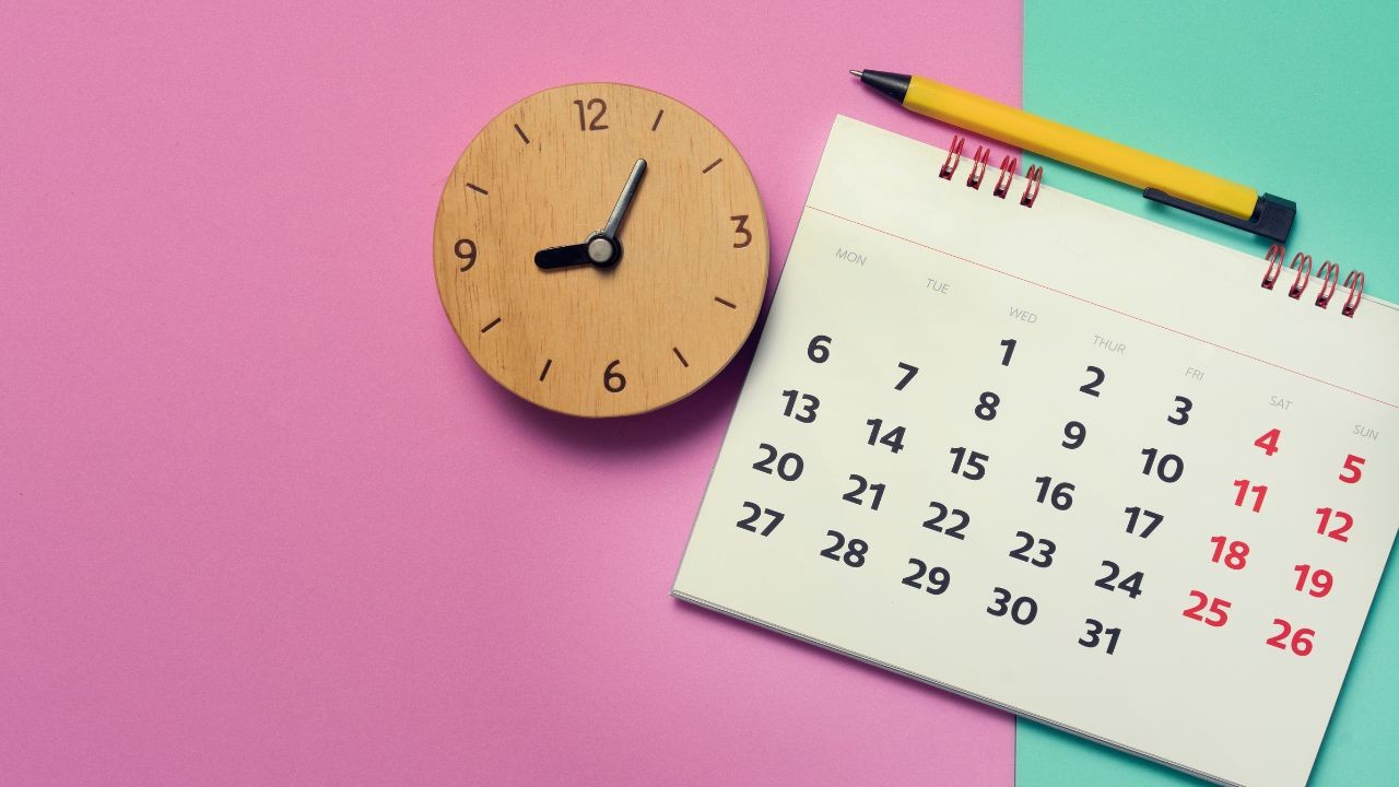 Relógio de madeira redondo marcando 8h10 e calendário de mesa com caneta amarela sobre fundo dividido em rosa e verde-água