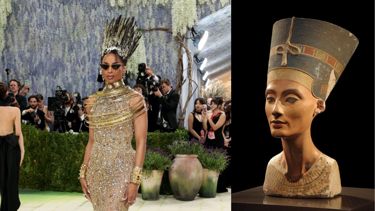 Uma pessoa com um vestido dourado ornamentado e um alto adorno de cabeça com pontas exibe-se com confiança em um evento glamoroso. Fotógrafos e plantas compõem o cenário ao fundo. Um busto de Nefertiti está exposto ao lado.