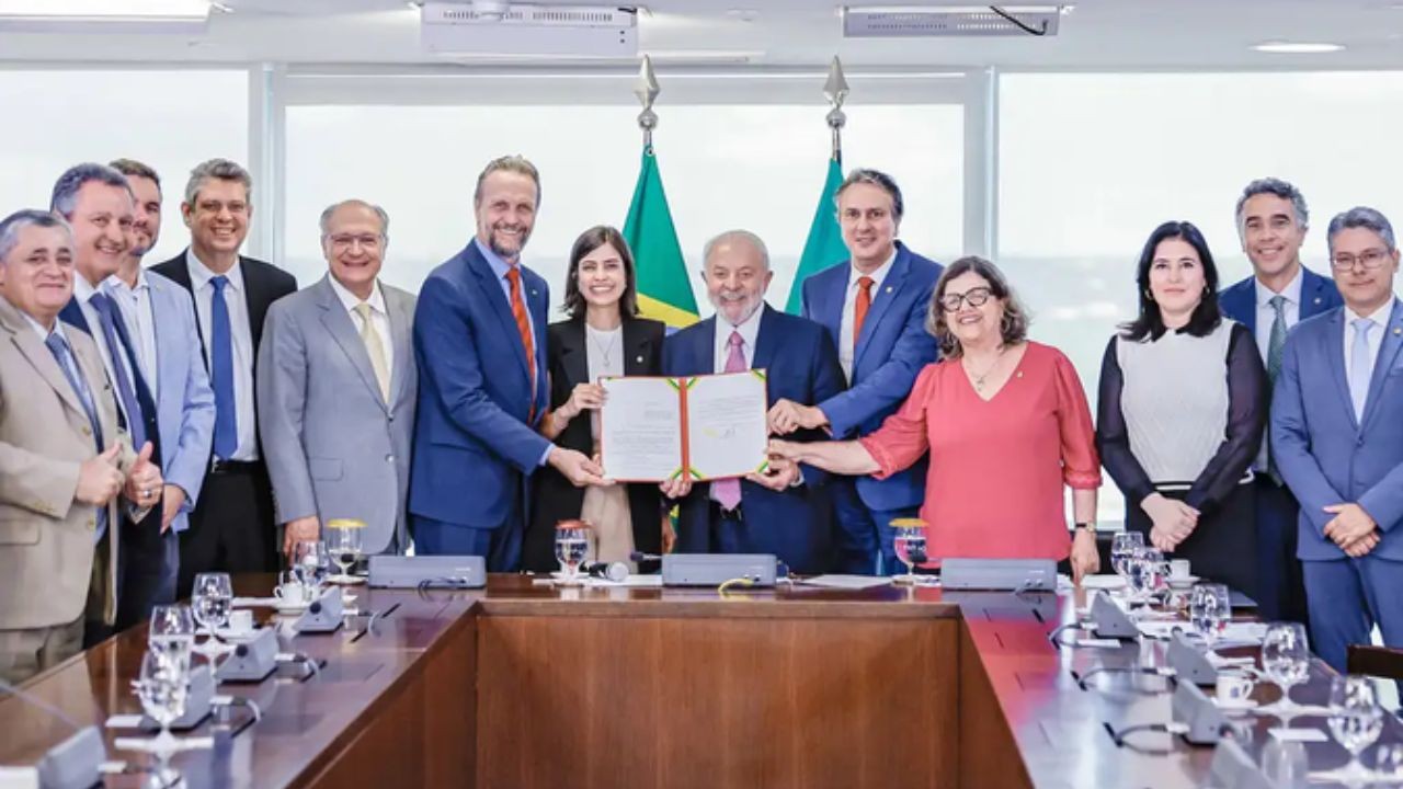 Lula e outras 13 pessoas, incluindo homens de terno e mulheres de blusas, sorriem enquanto assinam um documento em uma mesa de reunião, com bandeiras do Brasil ao fundo
