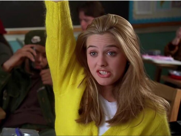 Uma adolescente com um suéter amarelo brilhante levanta a mão com entusiasmo em uma sala de aula. Outra aluna é visível ao fundo.