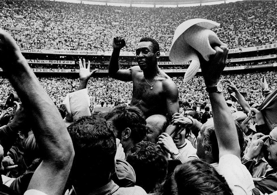 Pelé, sem camisa, é carregado nos ombros por torcedores em um estádio lotado, com o punho erguido em celebração. Ele usa um colar e sorri, enquanto a multidão ao redor levanta os braços, alguns segurando chapéus. O fundo mostra arquibancadas repletas de pessoas e anúncios publicitários