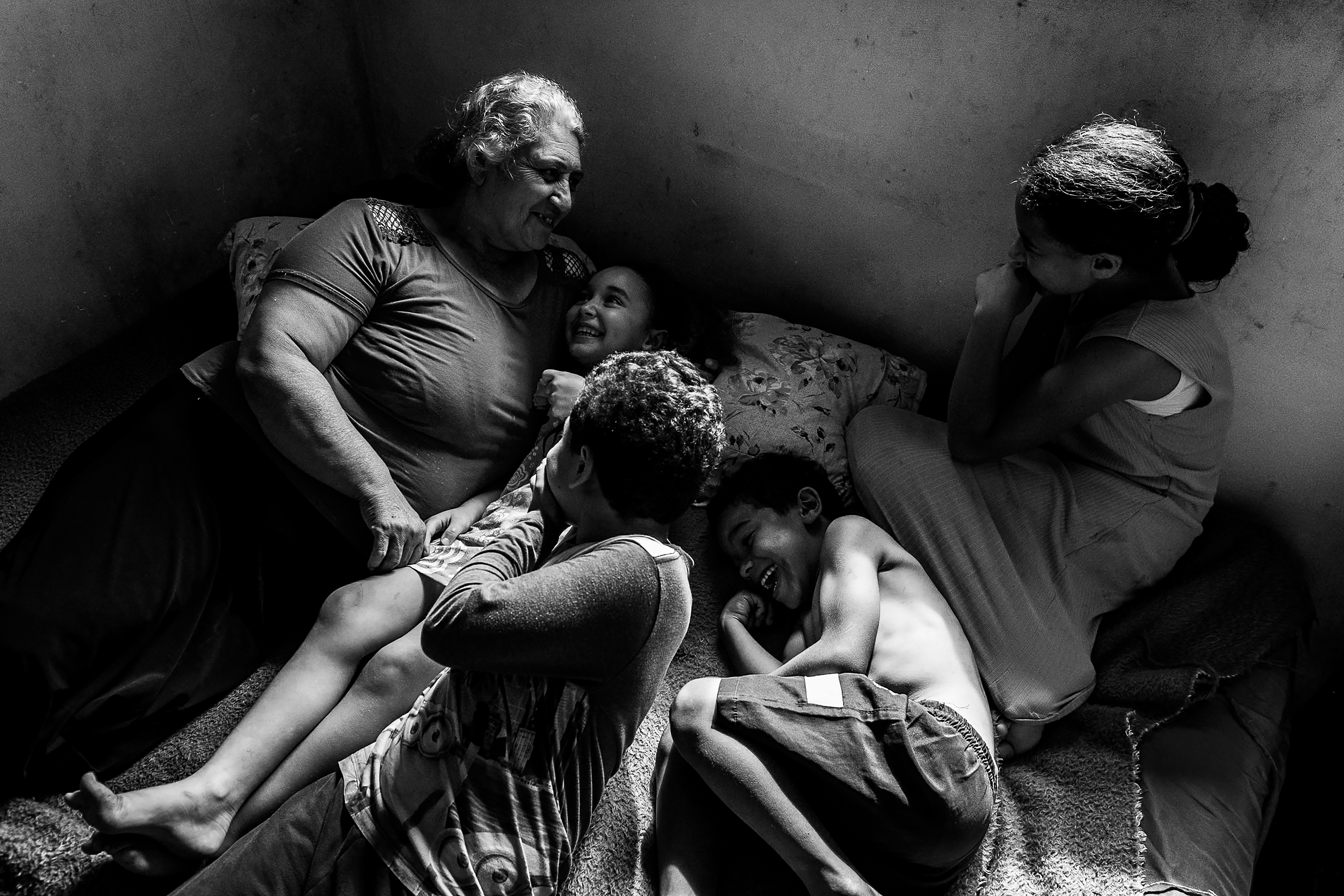 Fotografia em preto e branco de uma mulher idosa e quatro crianças, todos sorrindo e interagindo em um ambiente escuro. A mulher está sentada à esquerda, abraçando uma menina. Outras três crianças estão deitadas ou sentadas no chão, rindo. A luz incide sobre os rostos e corpos, criando um contraste dramático.