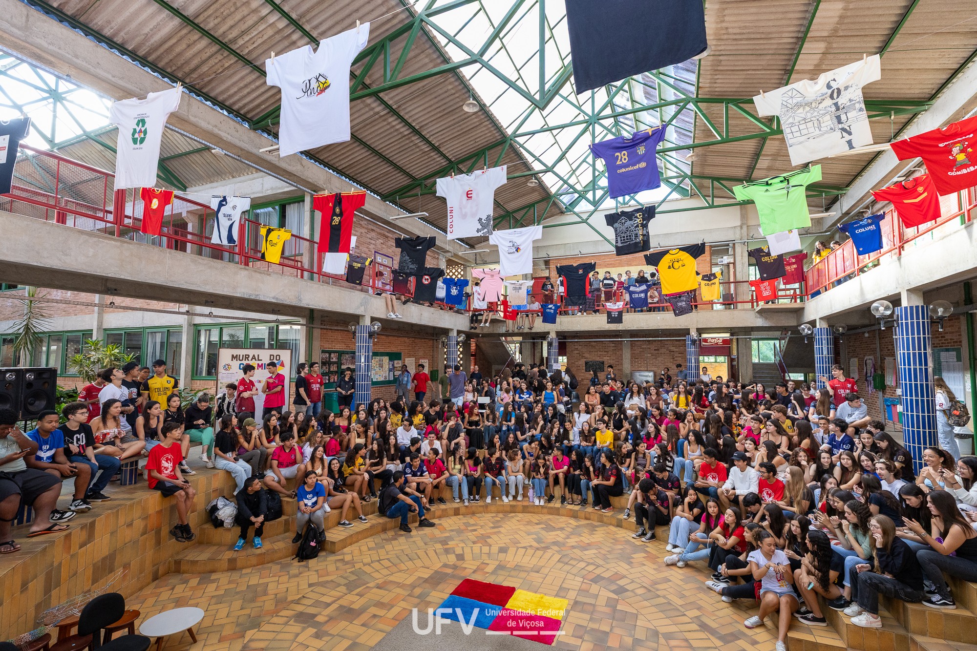 Grande grupo de jovens sentados em arquibancadas de madeira em um pátio interno, com camisetas coloridas penduradas no teto e na varanda superior. No chão, um logo da UFV