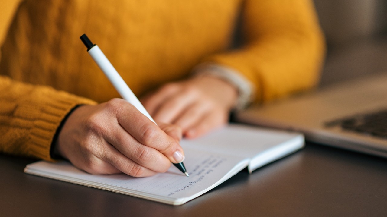 Pessoa com suéter mostarda escrevendo em um caderno aberto com caneta branca, ao lado de um notebook prateado em uma mesa escura