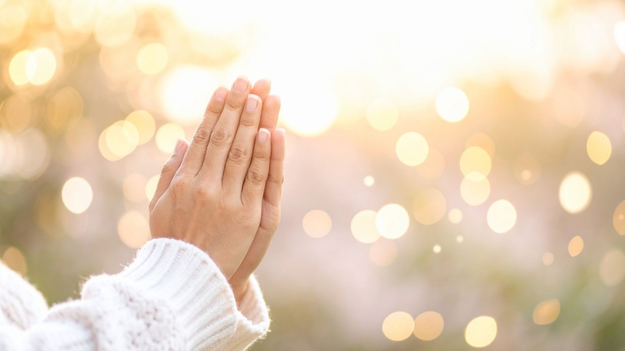 Mãos de uma pessoa com suéter branco em posição de oração, com fundo desfocado de luzes douradas e brancas, sugerindo um ambiente espiritual ou de esperança
