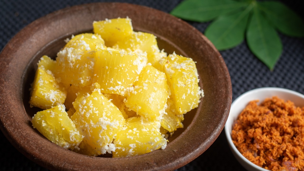 Mandioca cozida em pedaços amarelos, polvilhada com coco ralado branco, em uma tigela de barro marrom. Ao lado, uma tigela branca com farofa vermelha e folhas verdes ao fundo