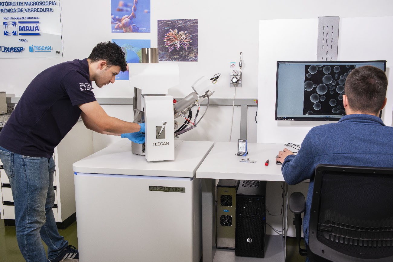 Dois homens em um laboratório. Um, de camiseta azul-marinho e jeans, usa luvas azuis para manusear um equipamento branco com a marca TESCAN. O outro, de blusa azul, está sentado de costas, operando um computador que exibe imagens de bolhas em preto e branco. Pôsteres científicos decoram a parede branca.