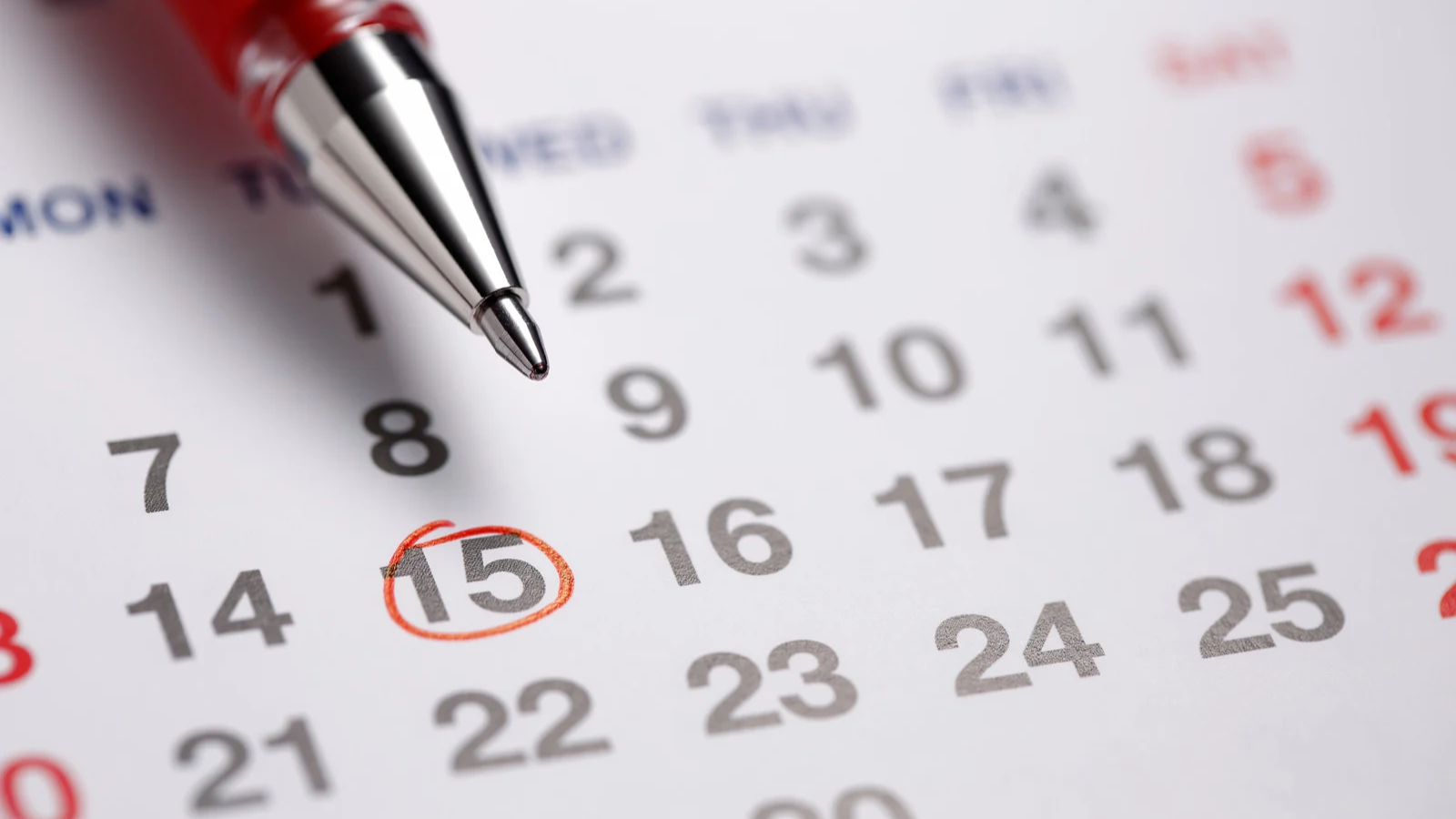 Ponta de uma caneta esferográfica vermelha e prateada sobre um calendário branco, com o número 15 circulado em vermelho, indicando uma data importante